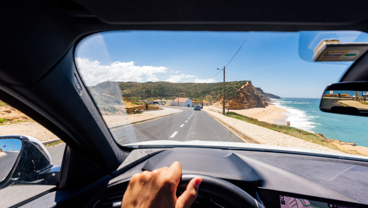 Blick über ein Lenkrad durch die Frontscheibe eines fahrenden Autos auf einer Straße an einer Küste mit Sandstrand bei Sonnenschein.