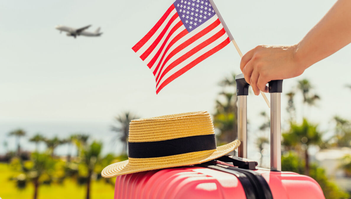 Ein Strohhut auf einem pinken Hartschalenkoffer, eine Hand am Griff hält eine kleine US-amerikanische Flagge, im Hintergrund Plamen und ein Flugzeug am Himmel.