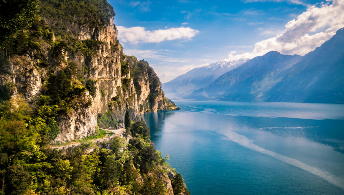 Bewachsene Felswand nahe Riva del Garda direkt am Gardasee mit Gipfeln auf der anderen Seeseite im Hintergrund.