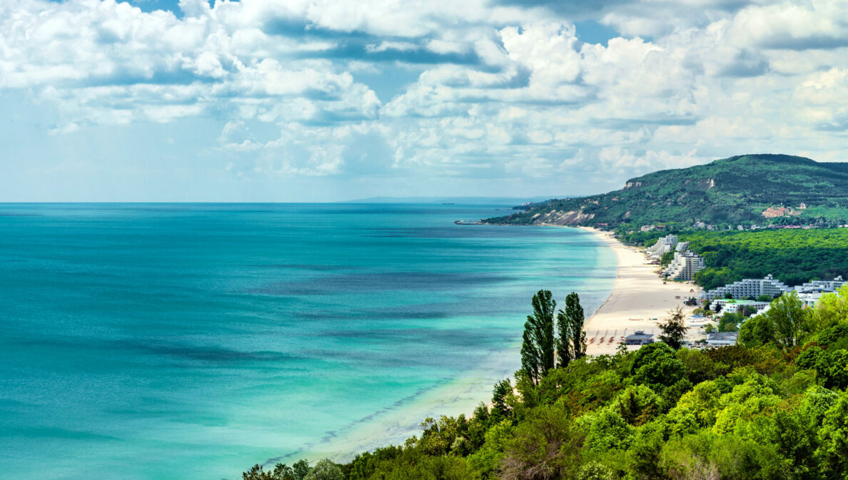 Grüner Küstenabschnitt mit Hotelanlagen an einem breiten Sandstrand vor flachem, türkisblauem Meer.