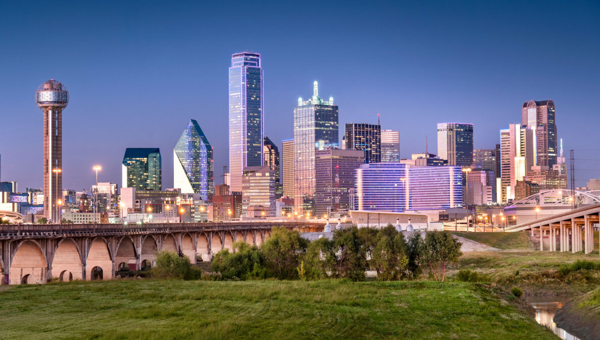 Skyline von Dallas mit Wolkenkratzern hinter einer Grünfläche mit Brückenbauten bei Abenddämmerung.