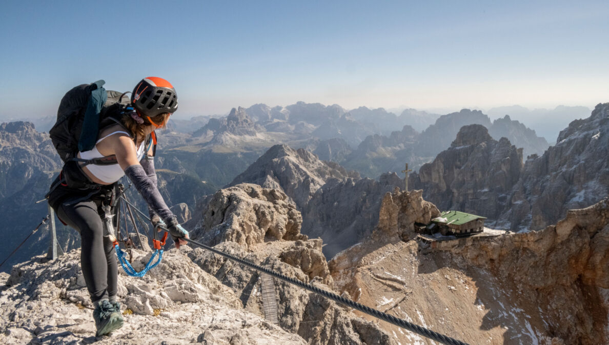 Eine Person mit Kletterausrüstung, die sich an einem Seil festhält, im Hintergrund Berge.