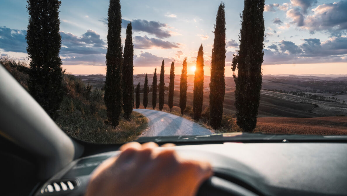 Blick auf eine mit Zypressen gesäumte Landstraße in Hügellandschaft bei Sonnenuntergang durch die Frontscheibe eines Autos, im Vordergrund eine Hand am Lenkrad.