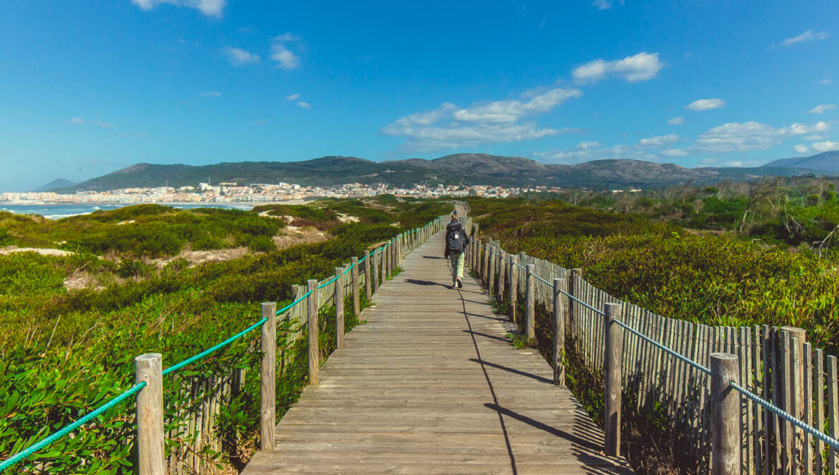 Eine unkenntliche Person beim Wandern in Portugal auf einem Holzsteg, der in Küstennähe über grüne Vegetation führt.