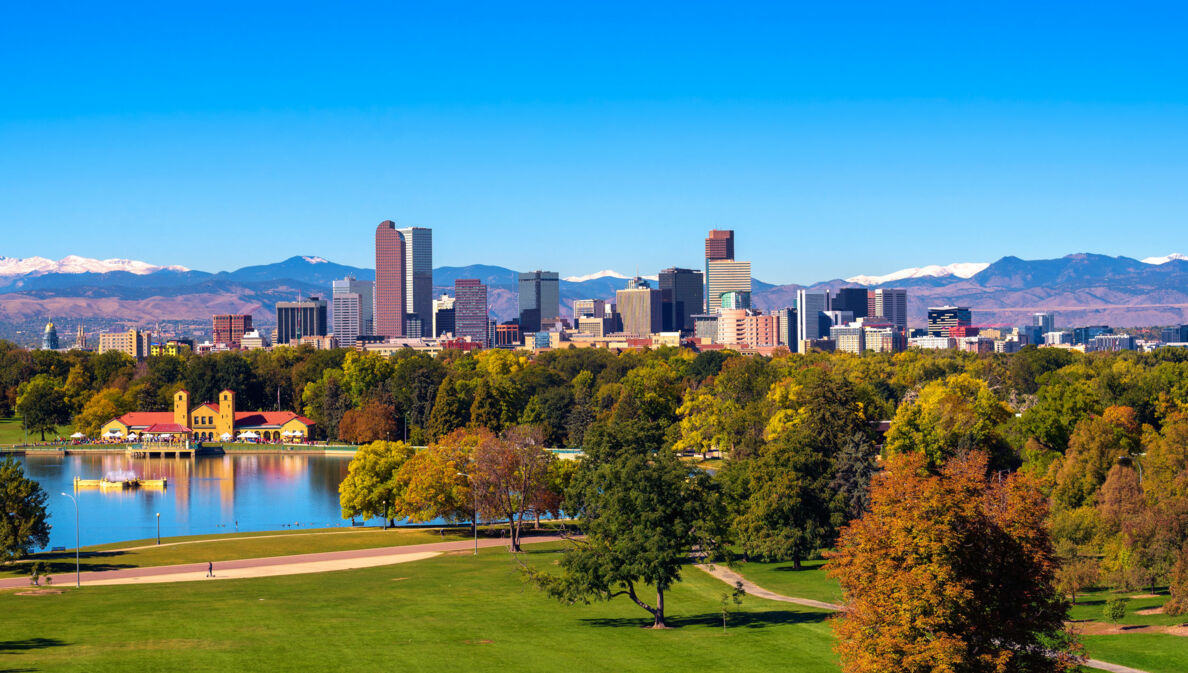 Skyline von Denver unter blauem Himmel vor Gebirgszug, im Vordergrund eine Parkanlage mit Teich und grünen Bäumen.