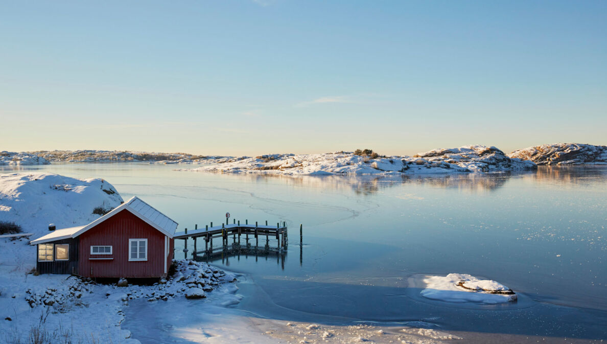 Ein rotes Holzhaus mit Steg an einem zugefrorenen See in verschneiter Landschaft in Schweden.