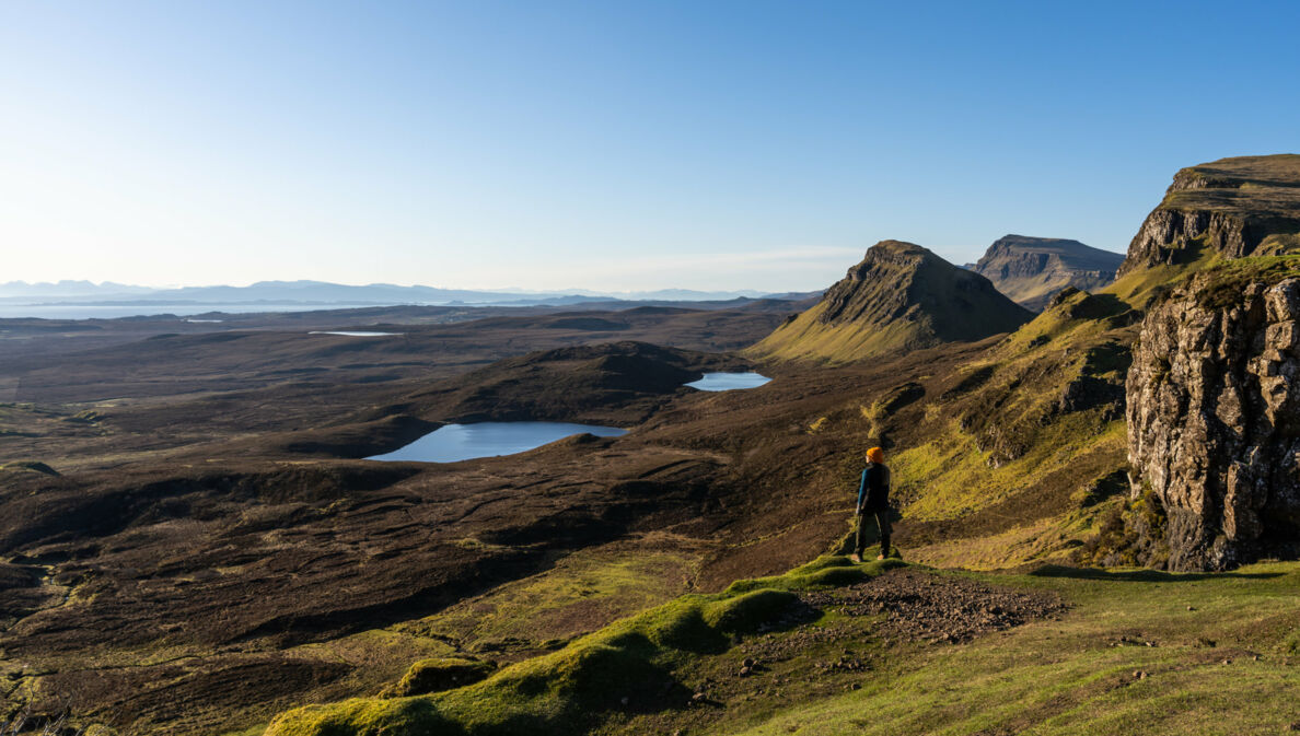 Eine Person steht in einer Landschaft aus Wiesen, Hügeln und Seen in Schottland. 