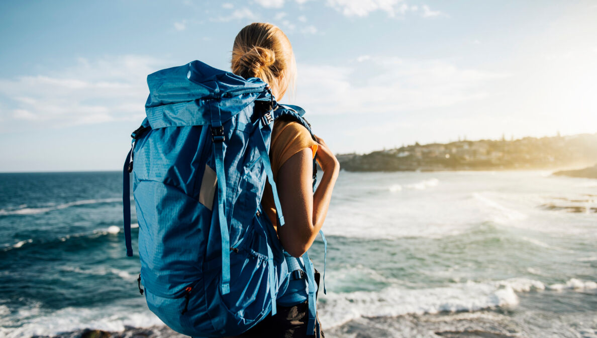 Rückansicht einer Frau mit großem blauen Rucksack am Strand, die aufs Meer blickt.