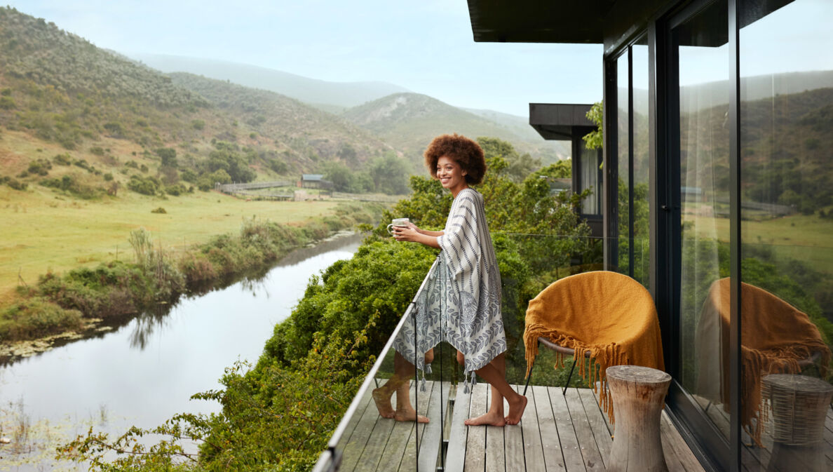 Eine lächelnde Frau steht mit einer Kaffeetasse auf dem Balkon eines Luxushotels mit Blick auf eine grüne Buschlandschaft mit Fluss.