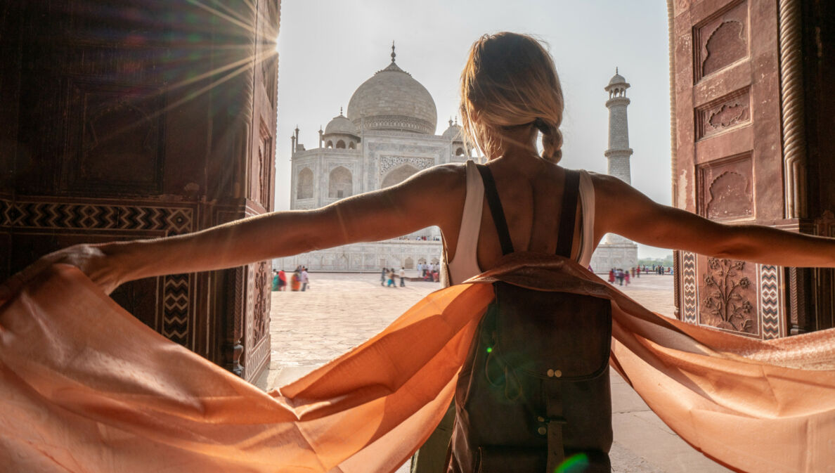 Rückansicht einer Frau mit Rucksack mit ausgestreckten Armen vor dem Taj Mahal.