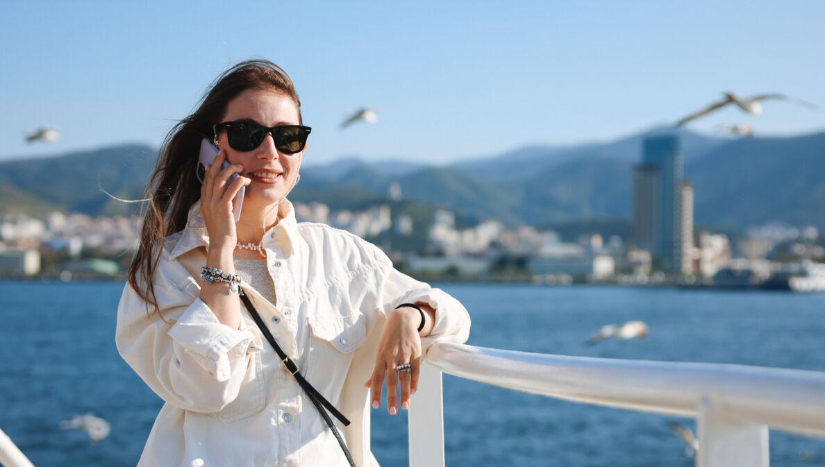 Eine Frau mit Sonnenbrille und wehenden Haaren telefoniert mit einem Smartphone an der Reling eines Schiffes auf dem Meer, im Hintergrund fliegen Möwen vor Stadtpanorama an einer Küste.