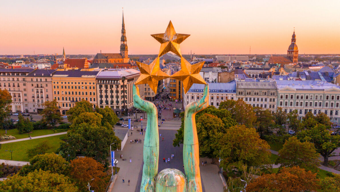 Stadtpanorama von Riga mit Altstadt aus der Luft, im Vordergrund die Arme einer Freiheitsstatue mit drei Sternen.