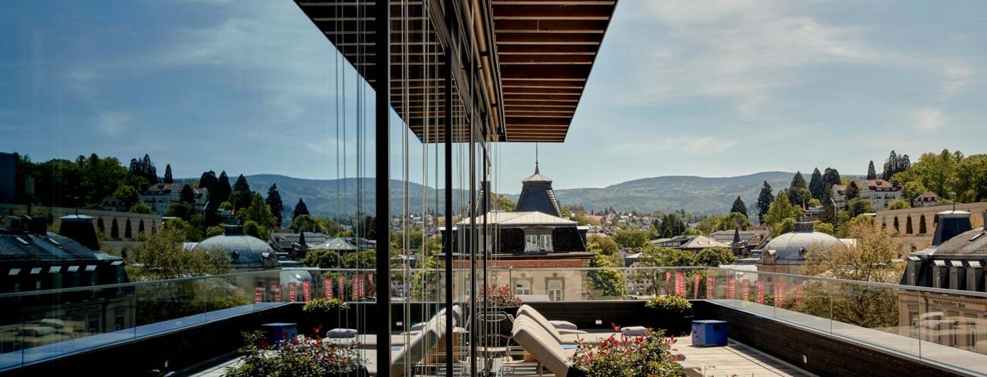 Große Balkonterrasse mit Liegestühlen vor grüner Hügellandschaft, die sich in der Glasfassade des Gebäudes spiegelt.