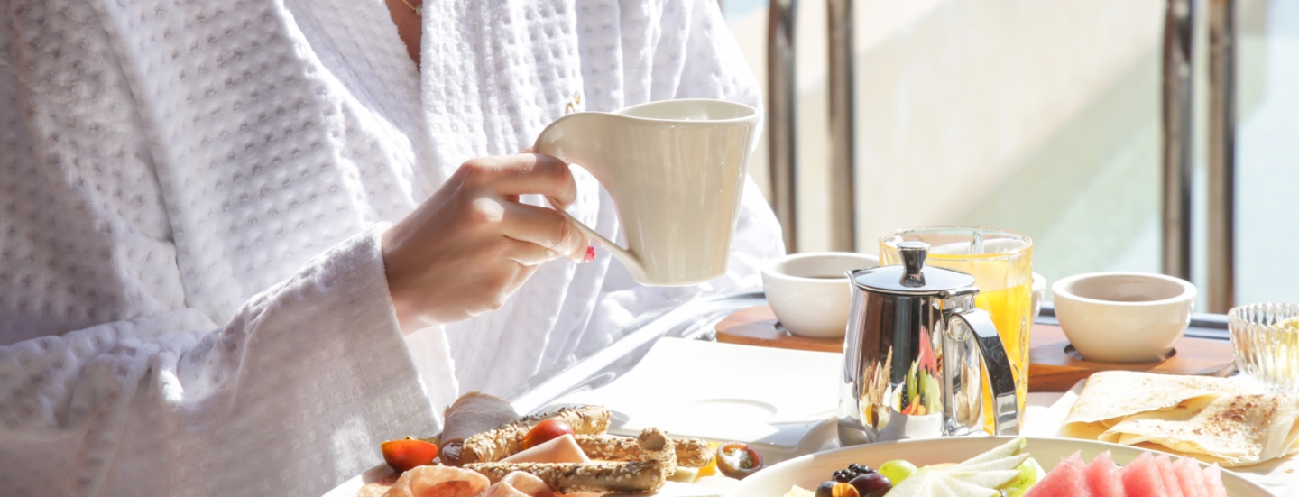 Eine Person sitzt an einem Tisch und hält eine Tasse in der Hand. Vor ihr steht ein Frühstückstablett mit Essen und Getränken.