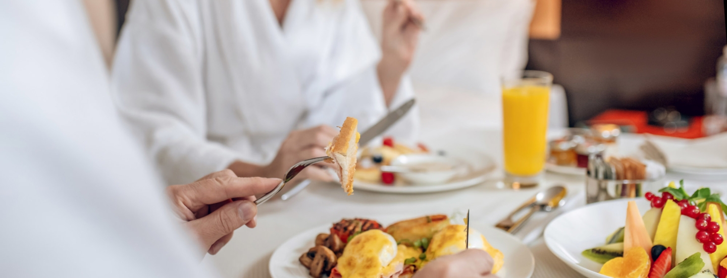 Nahaufnahme von zwei gesichtslosen Personen in weißen Bademänteln, die an einem eingedeckten Tisch in einem Hotelzimmer frühstücken.