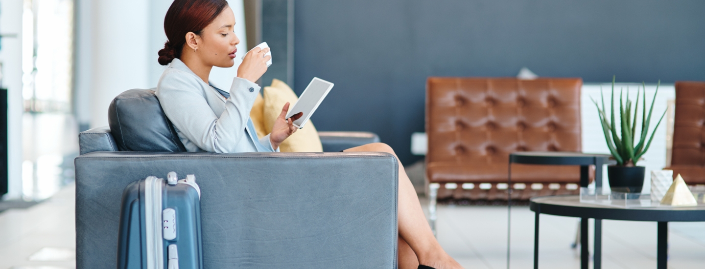 Eine Geschäftsfrau sitzt mit Tablet und einer Tasse Kaffee in den Händen im Sessel einer Lounge, daneben steht ein Trolley.