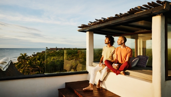 Zwei lässig gekleidete Männer sitzen auf einer Terrasse in der Abendsonne und schauen über eine tropische Landschaft aufs Meer