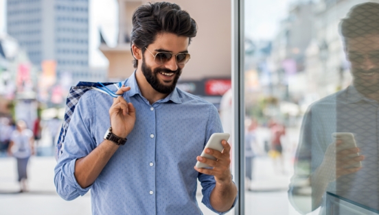 Ein junger Mann mit Vollbart, Sonnenbrille und Einkaufstüten in der Hand schaut in einer Einkaufsstraße lächelnd auf sein Smartphone.