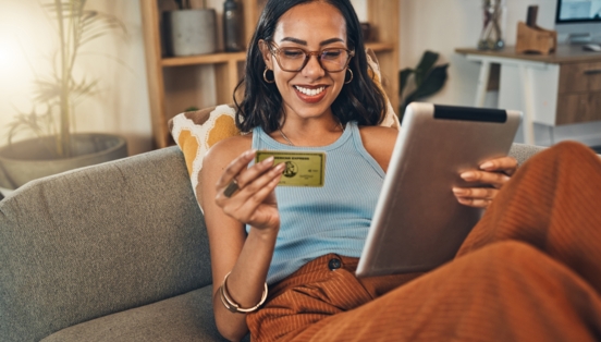 Eine junge Frau sitzt lächelnd auf einem Sofa, in ihren Händen hält sie ein Tablet und eine goldene Kreditkarte von American Express.