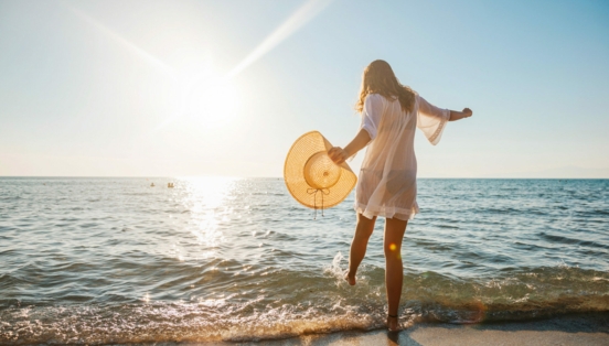 Eine junge Frau im weißen Sommerkleid und mit Strohhut in der Hand planscht mit ihrem Fuß im Wasser an einem Sandstrand am Meer bei Sonnenuntergang