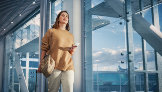 eine Frau am Flughafen, die mit einer Hand einen Koffergriff festhält und in der anderen Papiere hält.