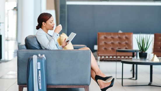 Eine Geschäftsfrau sitzt mit Tablet und einer Tasse Kaffee in den Händen im Sessel einer Lounge, daneben steht ein Trolley.