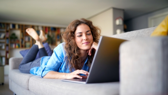 Eine Frau liegt bäuchlings auf einem Sofa in einem Wohnzimmer und schaut lächelnd auf einen Laptop vor ihr.