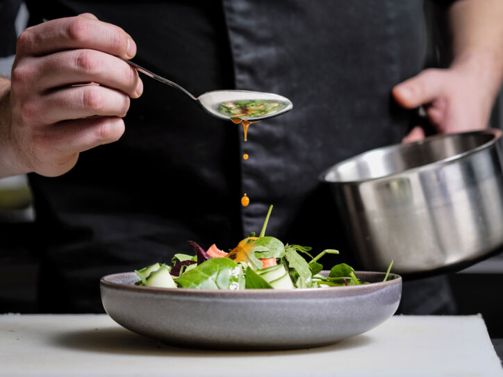 Nahaufnahme einer Hand, die mit einem Löffel gelbes Dressing über einen Teller mit Rohkost träufelt