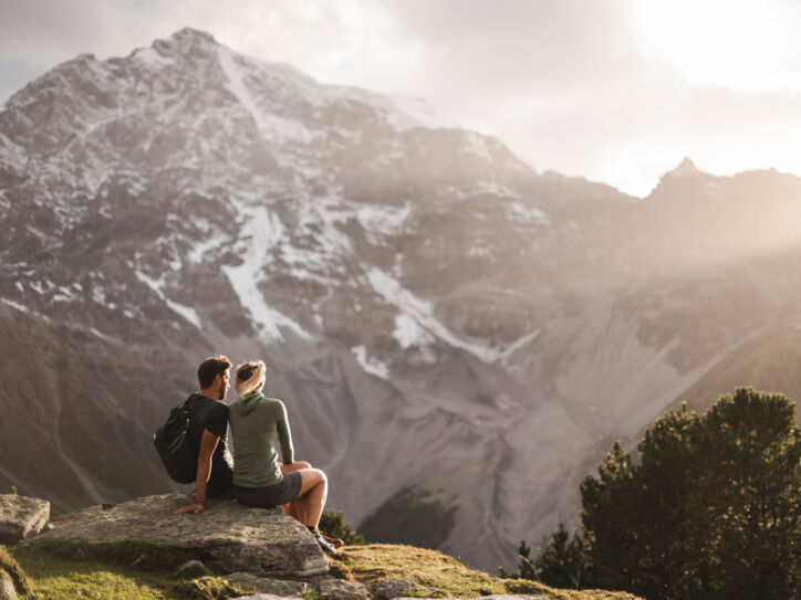 Zwei Personen sitzen im Nationalpark Stilfserjoch auf einem Stein und blicken auf ein Bergmassiv.