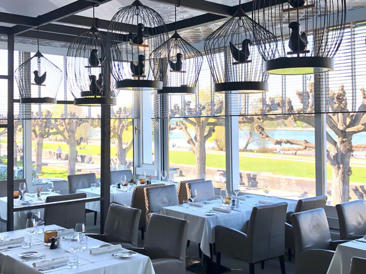 Speiseraum des Restaurants OX Royal mit eingedeckten Tischen und dekorativen Vogelkäfigen an der Decke in einem Wintergarten mit Panoramablick an der Uferpromenade eines Flusses.