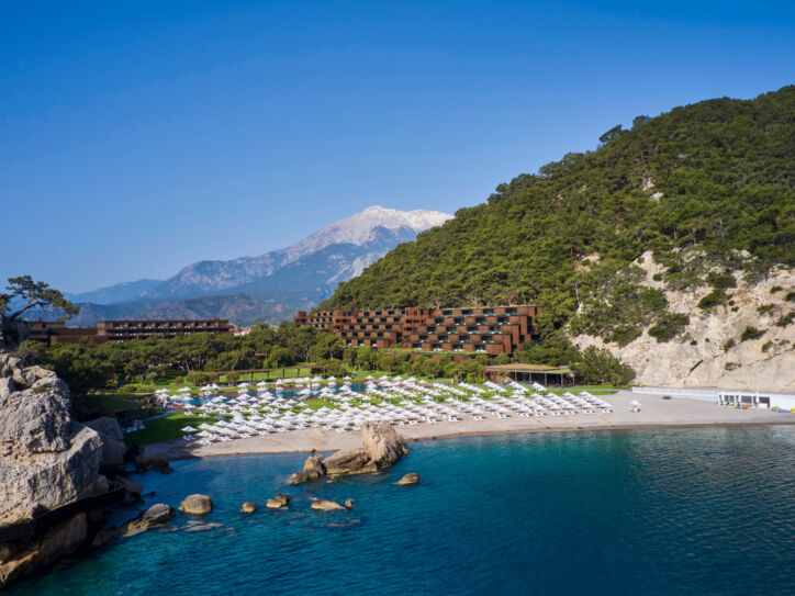 Ein Hotelresort in einer mediterranen Bucht mit Sandstrand, im Hintergrund eine Berglandschaft.