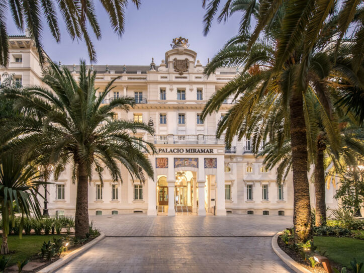 Imposante, helle Frontfassade eines Grandhotels mit Vorplatz, im Vordergrund Begrünung mit Palmen.