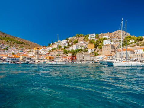 Mediterrane Ortschaft in Hügellage mit Hafenpromenade, im Vordergrund türkisblaues Meer mit Booten.