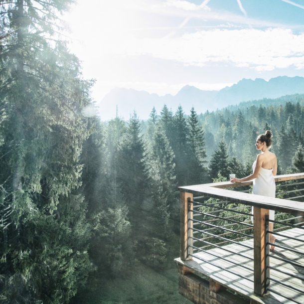 Eine in ein Handtuch gewickelte Frau blickt von einer Terrasse auf einen herrlichen Nadelwald