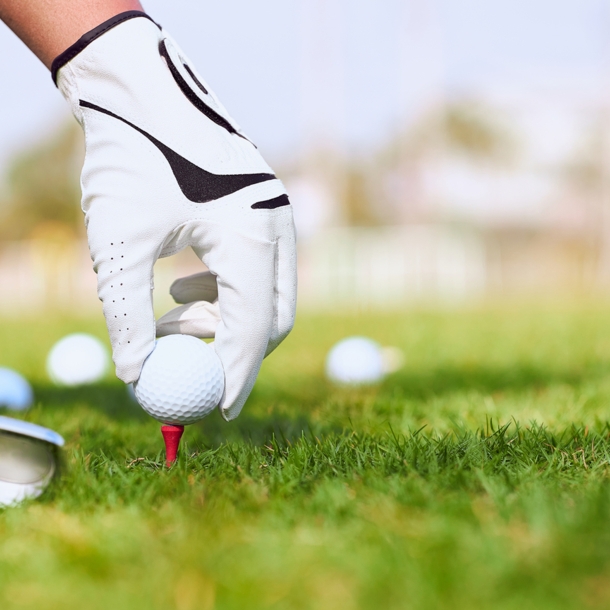 Detailaufnahme von Hand mit Handschuh, die Golfball auf Rasen platziert, daneben Golfschläger