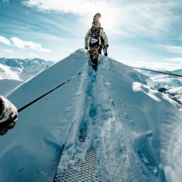 Ein:e Wintersportler:in mit Snowboard auf dem Rücken geht über einen schneebedeckten Berggipfel.