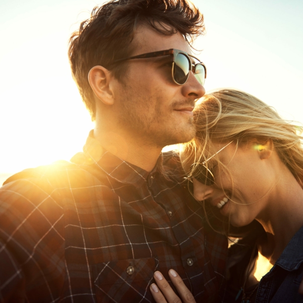 Eine Frau lehnt sich an ihren Partner, sie tragen beide eine Sonnenbrille, im Hintergrund sind das Meer und die untergehende Sonne zu erkennen