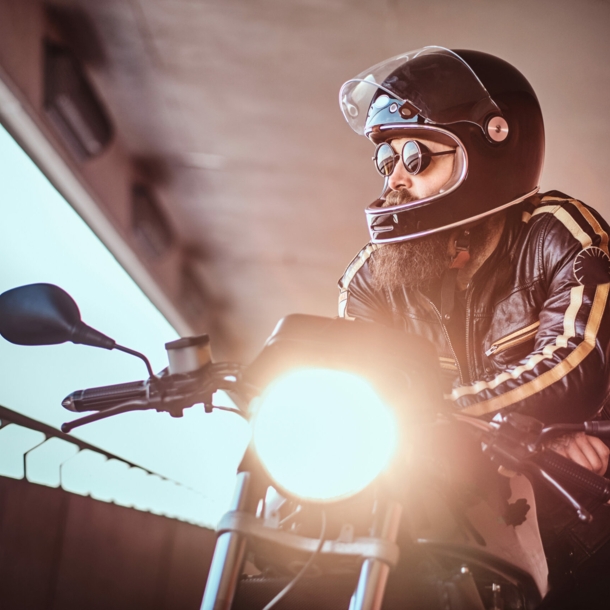 Ein junger, bärtiger Mann in Lederjacke lehnt lässig mit Helm und Sonnenbrille auf einem Motorrad und blickt aus einem offenen Betonbauwerk in die Ferne