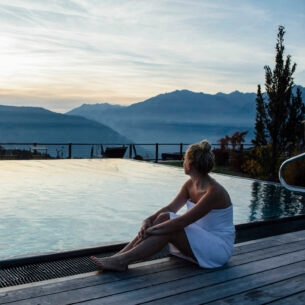 Eine Frau mit umwickelten Handtuch sitzt an einem Outdoor-Pool mit Blick auf die Berge in der Dämmerung