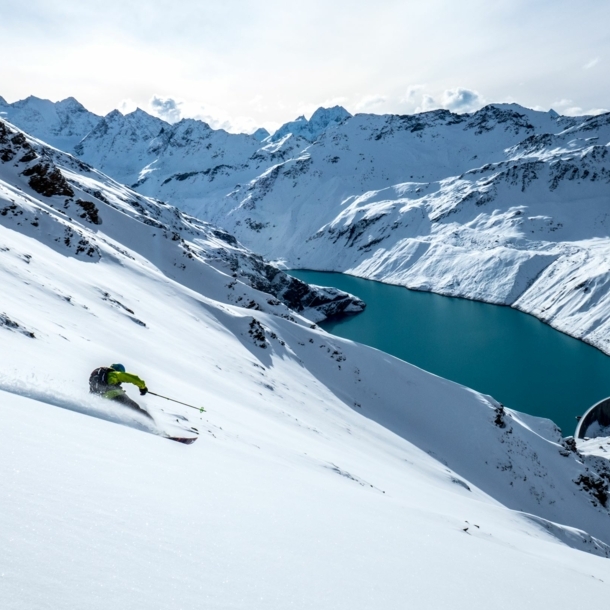 Eine Person auf Ski fährt eine Piste hinunter, im Hintergrund eine Bergkulisse.