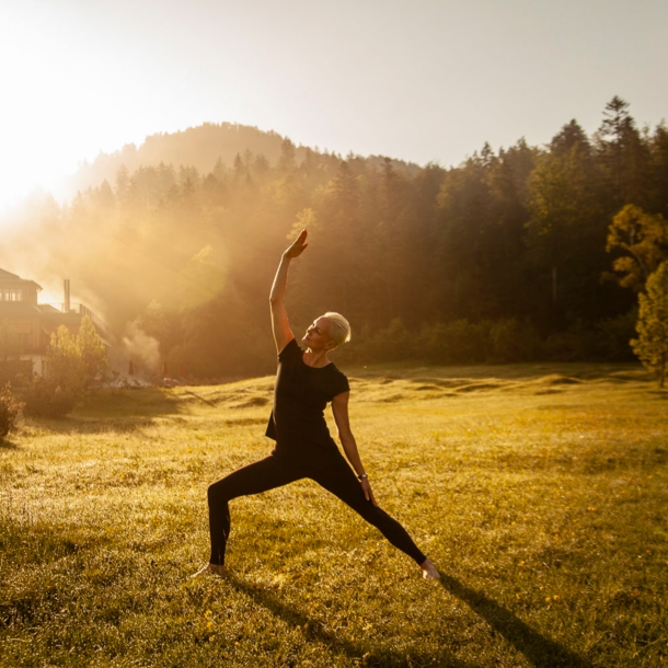 Eine Frau macht Yoga bei Sonnenaufgang und noch leichtem Nebel in einem grünen Garten