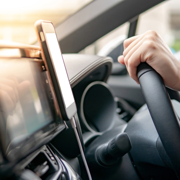 Ein Smartphone in einer Halterung am Armaturenbrett eines Autos, daneben zwei Hände an einem Lenkrad