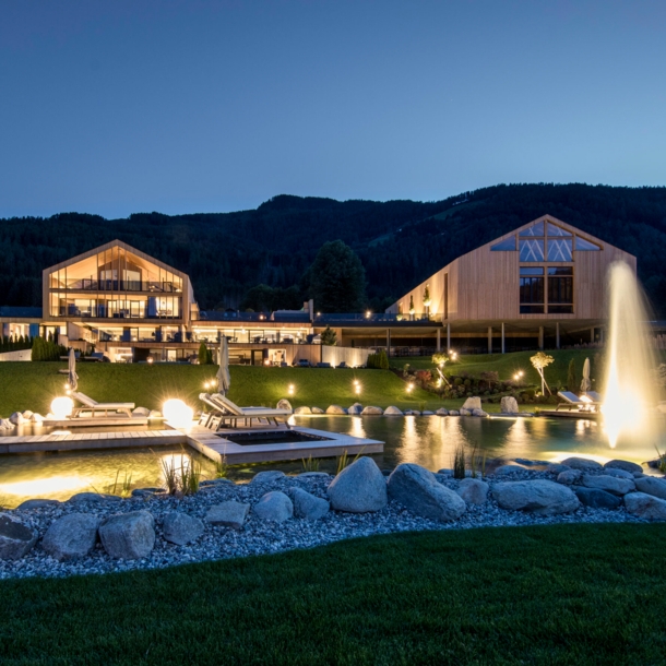 Eine luxuriöse, beleuchtete Hotelanlage aus Holzgebäuden mit Landschaftsgarten und Badeteich vor Bergpanorama bei Nacht