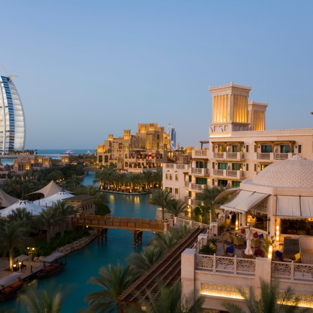 Stadtteil von Dubai mit Gebäuden an einem Fluss in der Abenddämmerung, im Hintergrund das Hotel Burj Al Arab in Segelform am Wasser