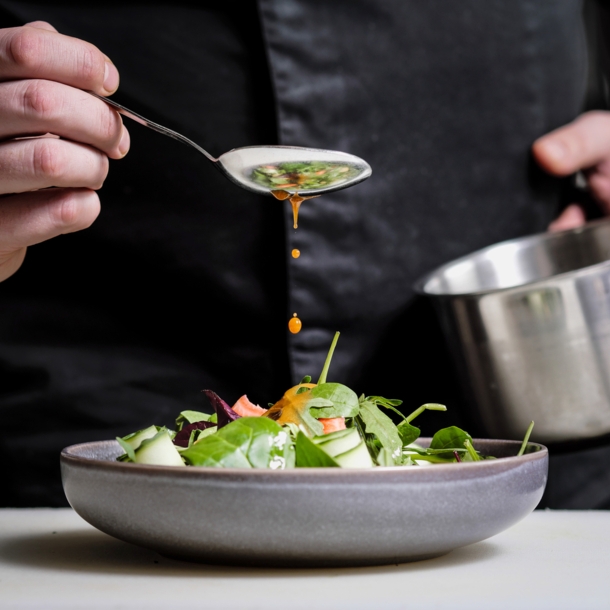 Nahaufnahme einer Hand, die mit einem Löffel gelbes Dressing über einen Teller mit Rohkost träufelt