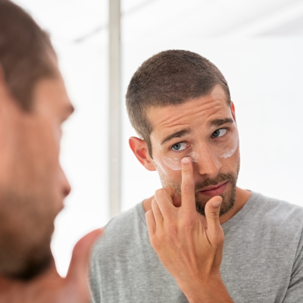 Ein Mann blickt in den Spiegel, während er sich unter den Augen Creme aufträgt