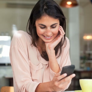 Eine junge Frau an einem Tisch in einem Café schaut lächelnd auf ihr Smartphone