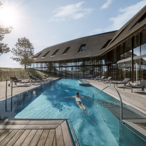 Ein luxuriöses Hotel in den Dünen mit Reetdach und Außenpool, in dem eine Frau schwimmt