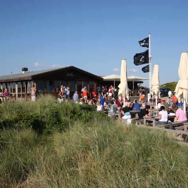 Viele Menschen sitzen an Holztischen und -bänken im Freien vor einem Haus.