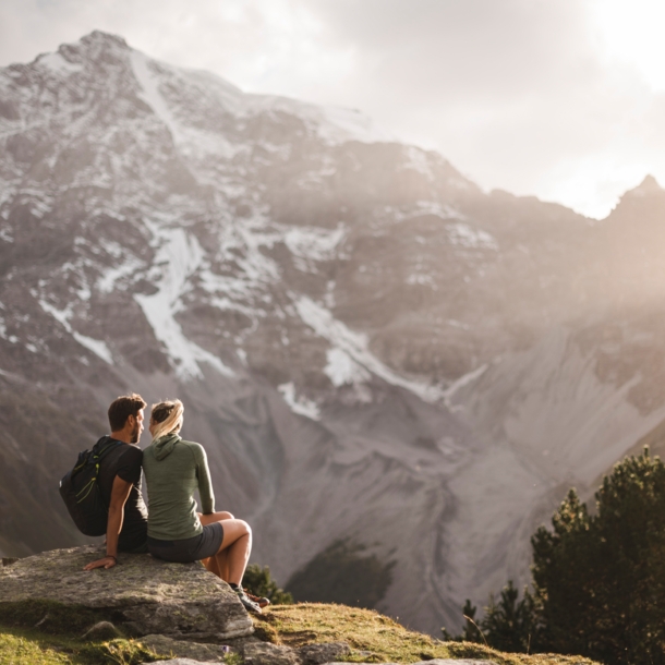 Zwei Personen sitzen im Nationalpark Stilfserjoch auf einem Stein und blicken auf ein Bergmassiv.